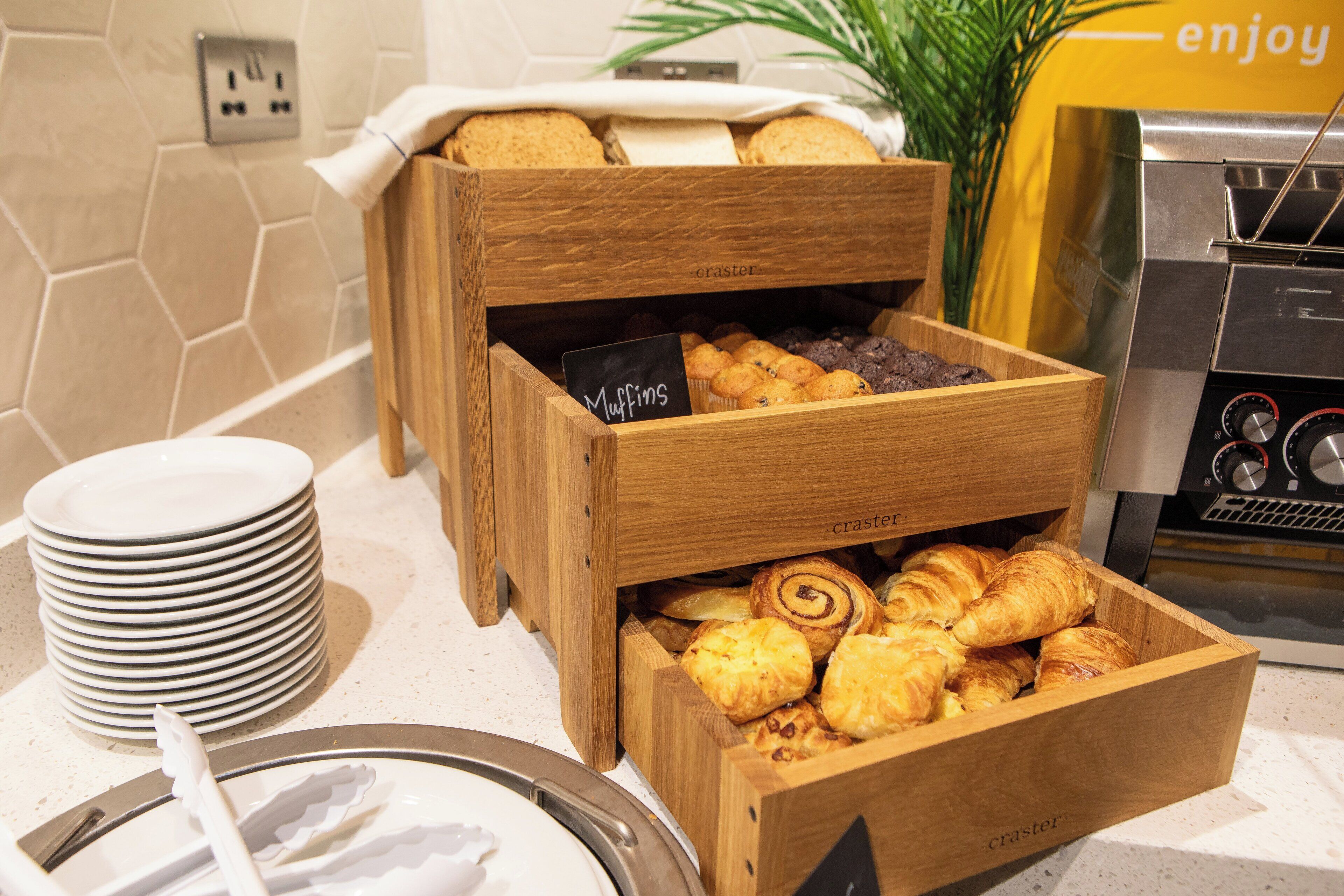 A breakfast buffet with wooden boxes containing muffins and pastries, a stack of white plates, and a coffee machine.