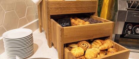 A breakfast buffet with wooden boxes containing muffins and pastries, a stack of white plates, and a coffee machine.
