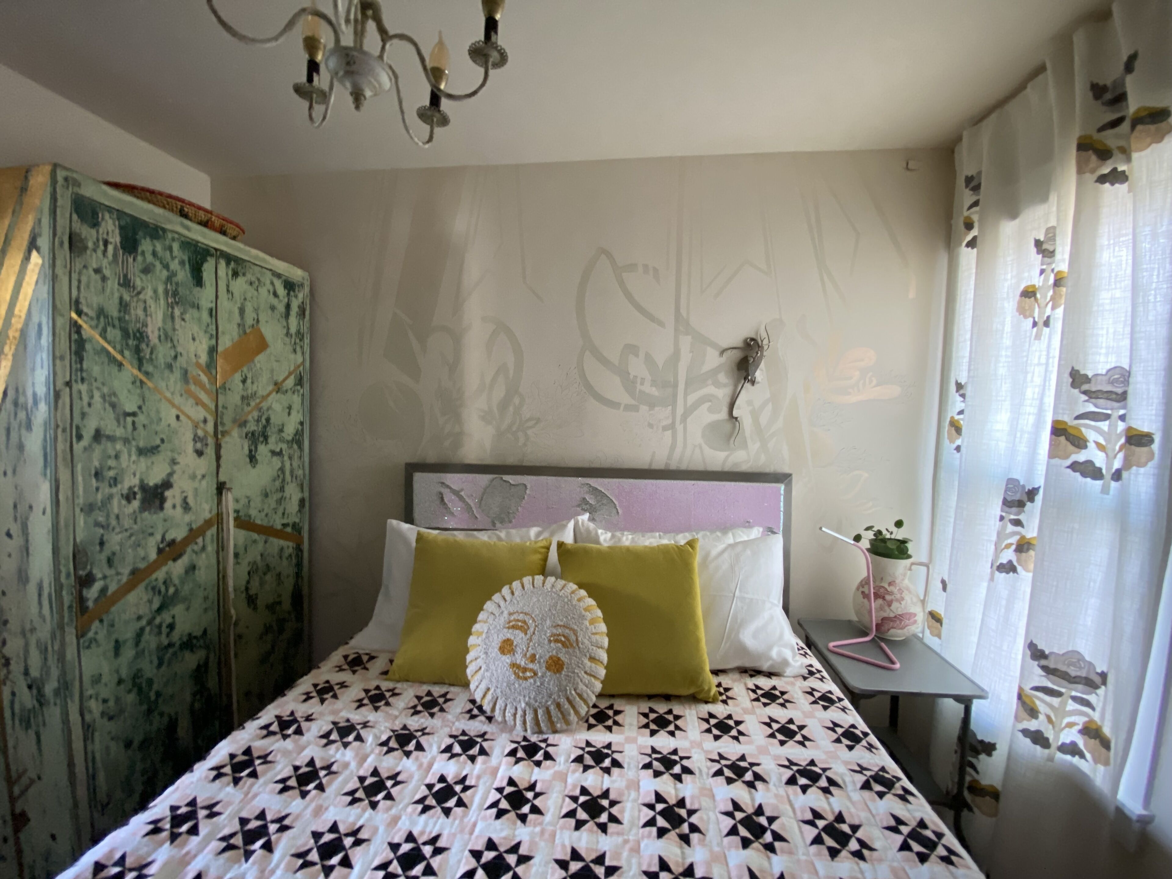 A bedroom with a patterned bedspread, a decorative headboard, a chandelier, a floral curtain, and a vintage wardrobe.