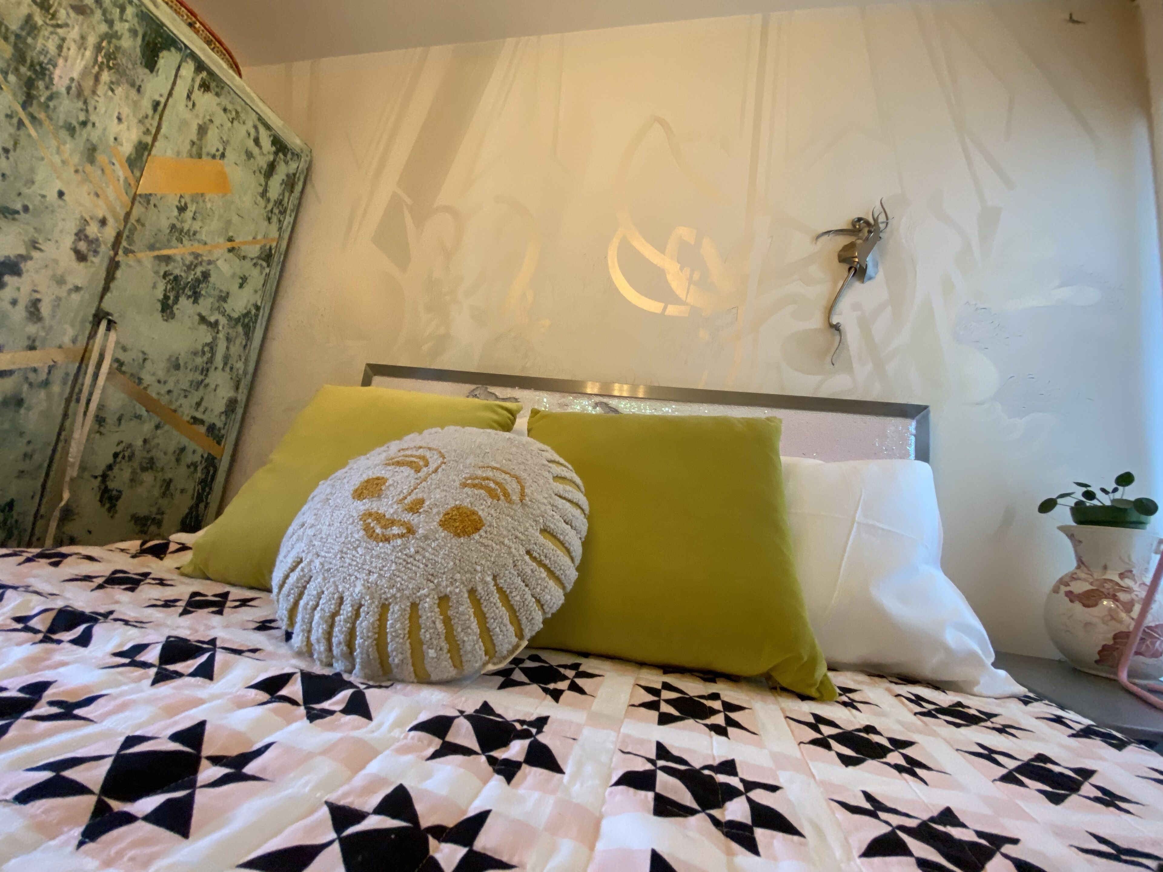 Un lit recouvert d’une couvre-lit à motifs géométriques, deux oreillers, dont l’un est orné d’un motif représentant un visage, et un mur avec un luminaire et une petite plante.