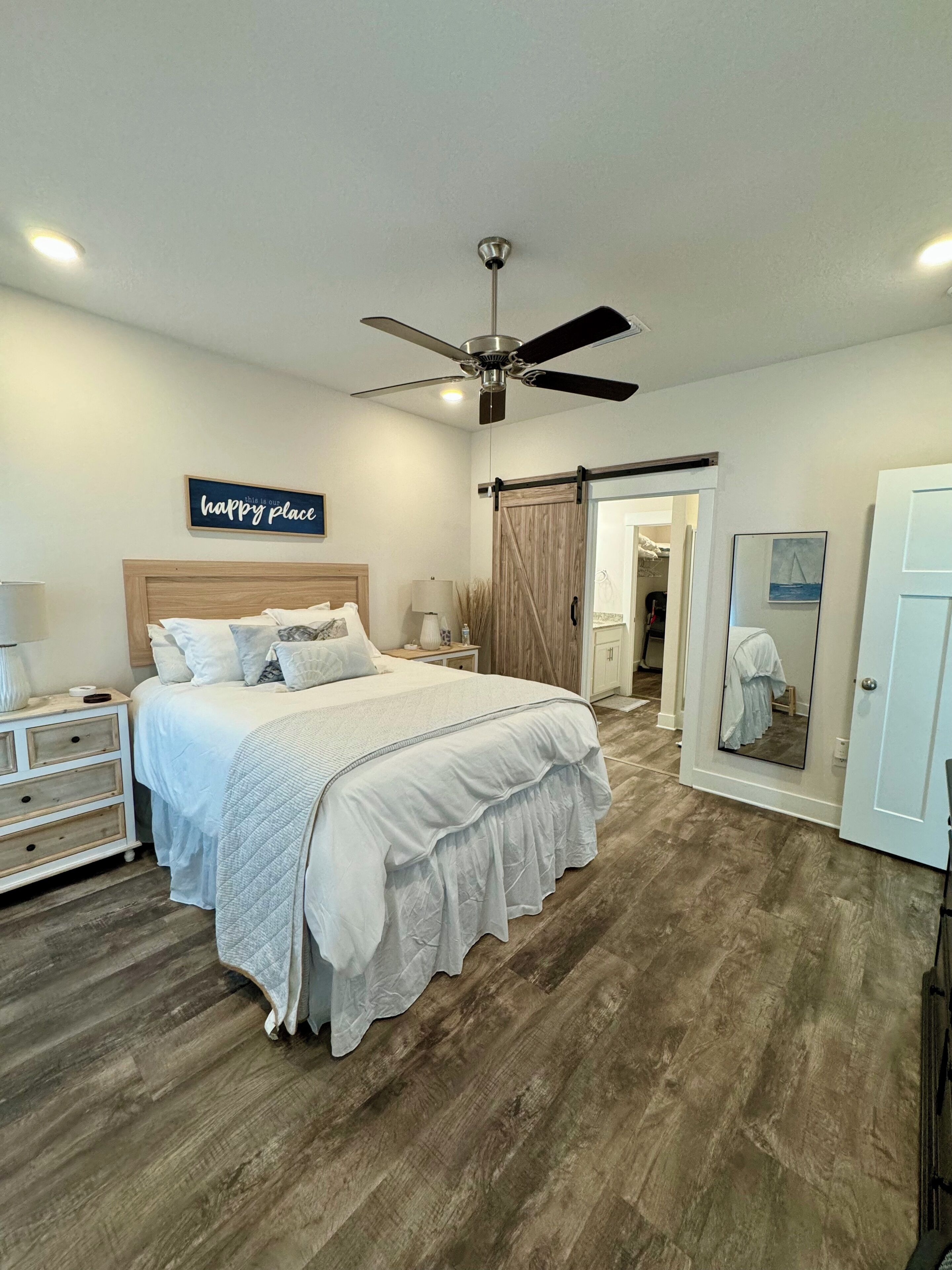 A bedroom with a bed, a ceiling fan, a sliding barn door, a mirror, and a 'happy place' sign on the wall.