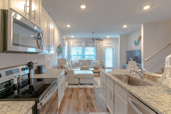 A modern kitchen with a built-in microwave, stove, and sink. There is a white sectional sofa in the living area, and a ceiling fan above.