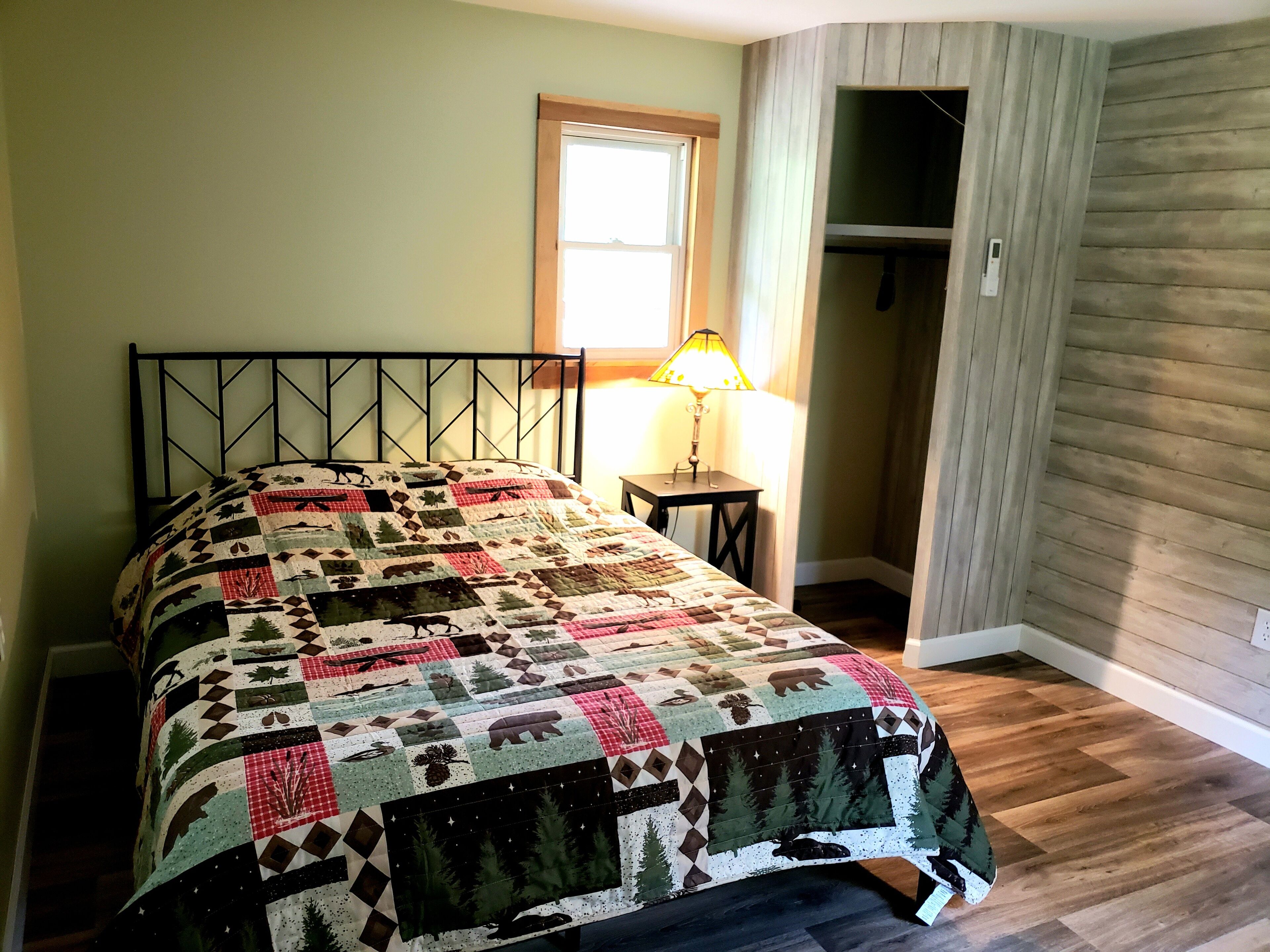 A bed with a patterned quilt, a bedside table with a lamp, and a closet in a room with wooden flooring and a window.