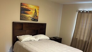 Un lit bien fait, avec une tête de lit en bois, un tableau représentant un voilier accroché au mur et un rideau à la fenêtre.