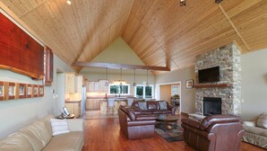 A spacious living area with a high ceiling, wooden beams, a stone fireplace, and a kitchen in the background. - The Bunkhouse Retreat /Sleeps 15/7BR/5BA/4 Private Acres (Kings Mountain)