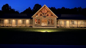 A two-story lodge with a prominent wooden gable roof and stone facade, featuring multiple windows and a well-lit entrance. - The Bunkhouse Retreat /Sleeps 15/7BR/5BA/4 Private Acres (Kings Mountain)