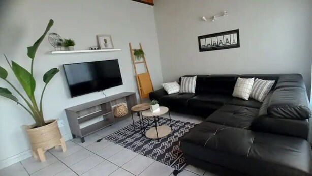 A living room with a black leather sofa, a flat-screen TV mounted on the wall, a small round table with a plant, and a patterned rug. - Lovely 1 bedroom Loft in the Heart of Walvisbay. (Walvis Bay)