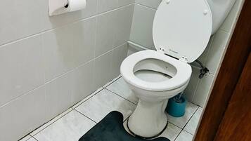 Un inodoro con tapa y asiento blancos, un dispensador de papel higiénico en la pared y un cubo azul de limpieza en el suelo.