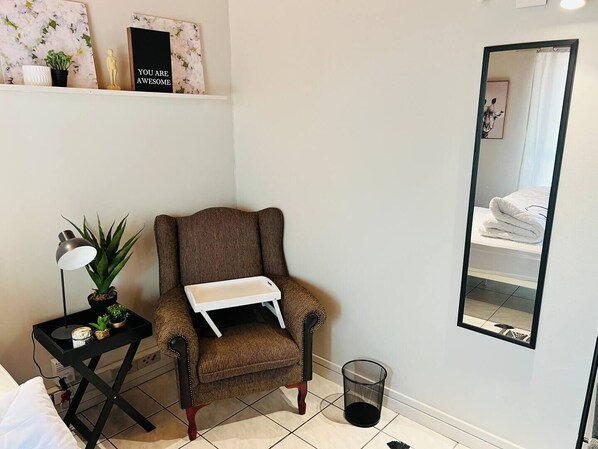 A room with a brown armchair, a small table with a lamp and plants, a mirror, and a shelf with decorative items.