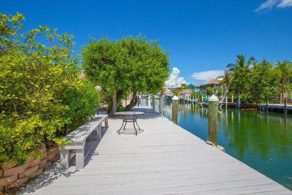 A wooden dock with a table and benches, overlooking a calm water body with boats and buildings in the background.
