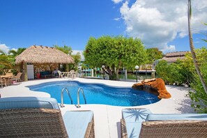 A swimming pool with lounge chairs, a thatched-roof structure, and lush greenery.