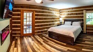 Ein Schlafzimmer in einer Blockhütte mit Holzboden, einem Bett mit weißer Bettwäsche, einem an der Wand befestigten Fernseher und einem Fenster mit Blick auf Grünflächen.
