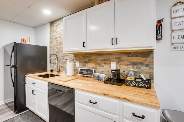 Cocina moderna con armarios blancos, refrigerador negro, encimera de madera y cartel de barra de café.