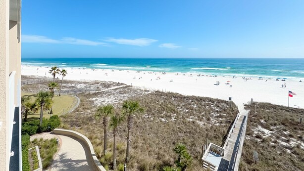 Blick auf einen Strand mit einem hölzernen Promenadenweg, Palmen und einem Sandstrand mit Menschen und Sonnenschirmen.