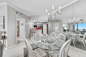 A modern dining area with a glass-top table surrounded by white chairs, a chandelier, and a view of the kitchen in the background. - BEACHFRONT CONDO, MULTIPLE POOLS, PRIVATE BALCONY! (Orange Beach)