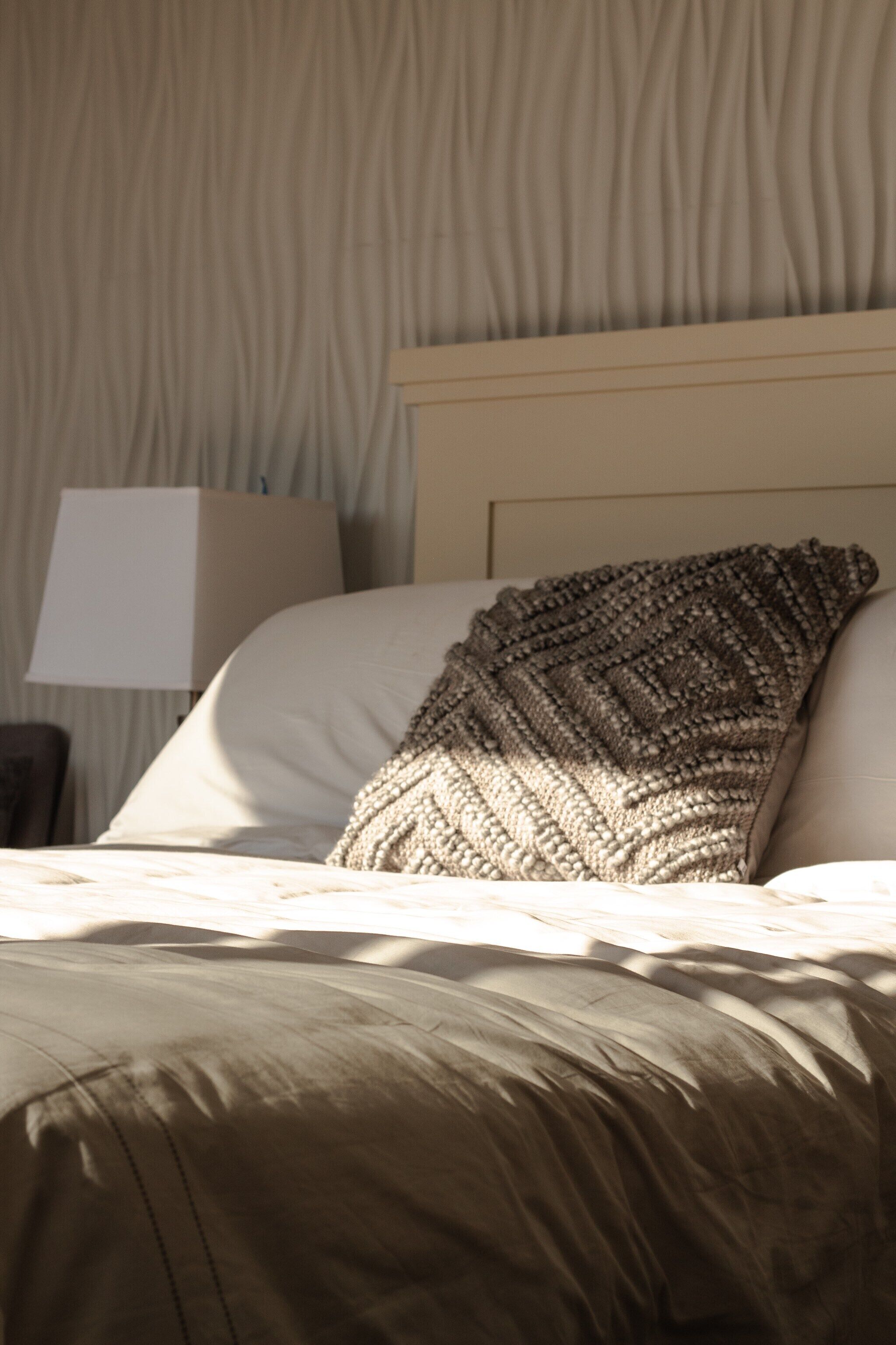 Una cama con una colcha texturizada y un almohadón, una lámpara y una pared con un diseño ondulado.
