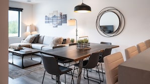 A modern living room with a dining area, featuring a sofa, coffee table, and a large wooden dining table with chairs. - Revylation by Revelstoke Vacations (Revelstoke)