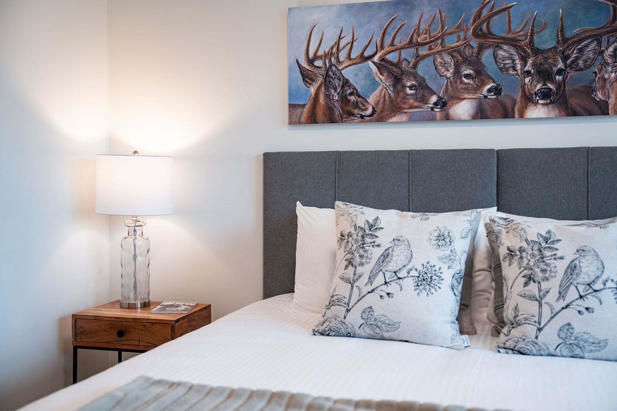 A neatly made bed with floral pillows, a bedside lamp, and a painting of deer on the wall.