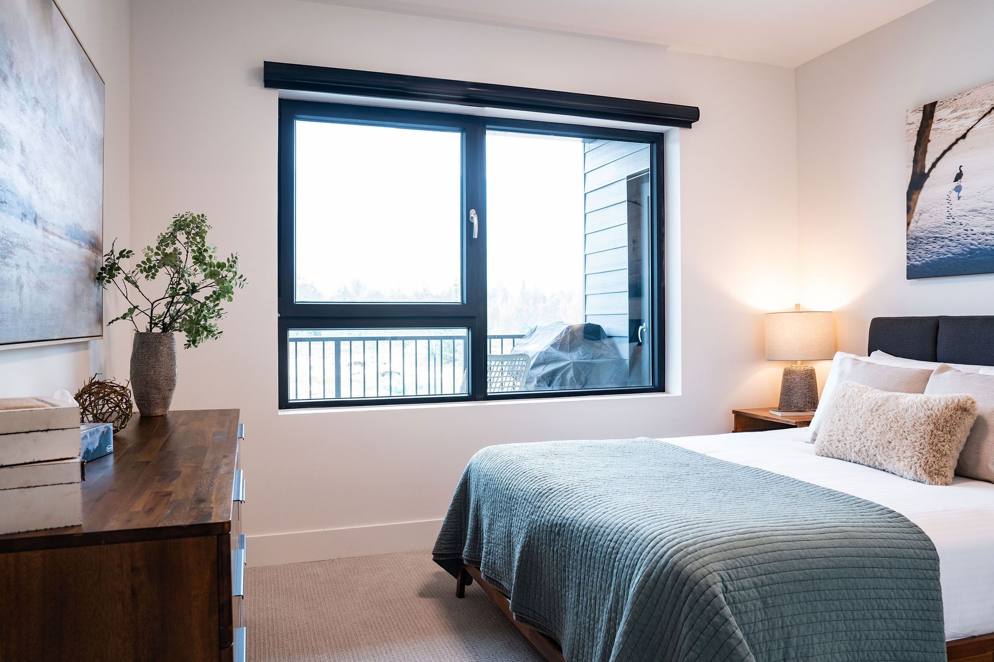 A bedroom with a large window, a bed with a green quilt, a bedside lamp, and a vase with a plant on a dresser.
