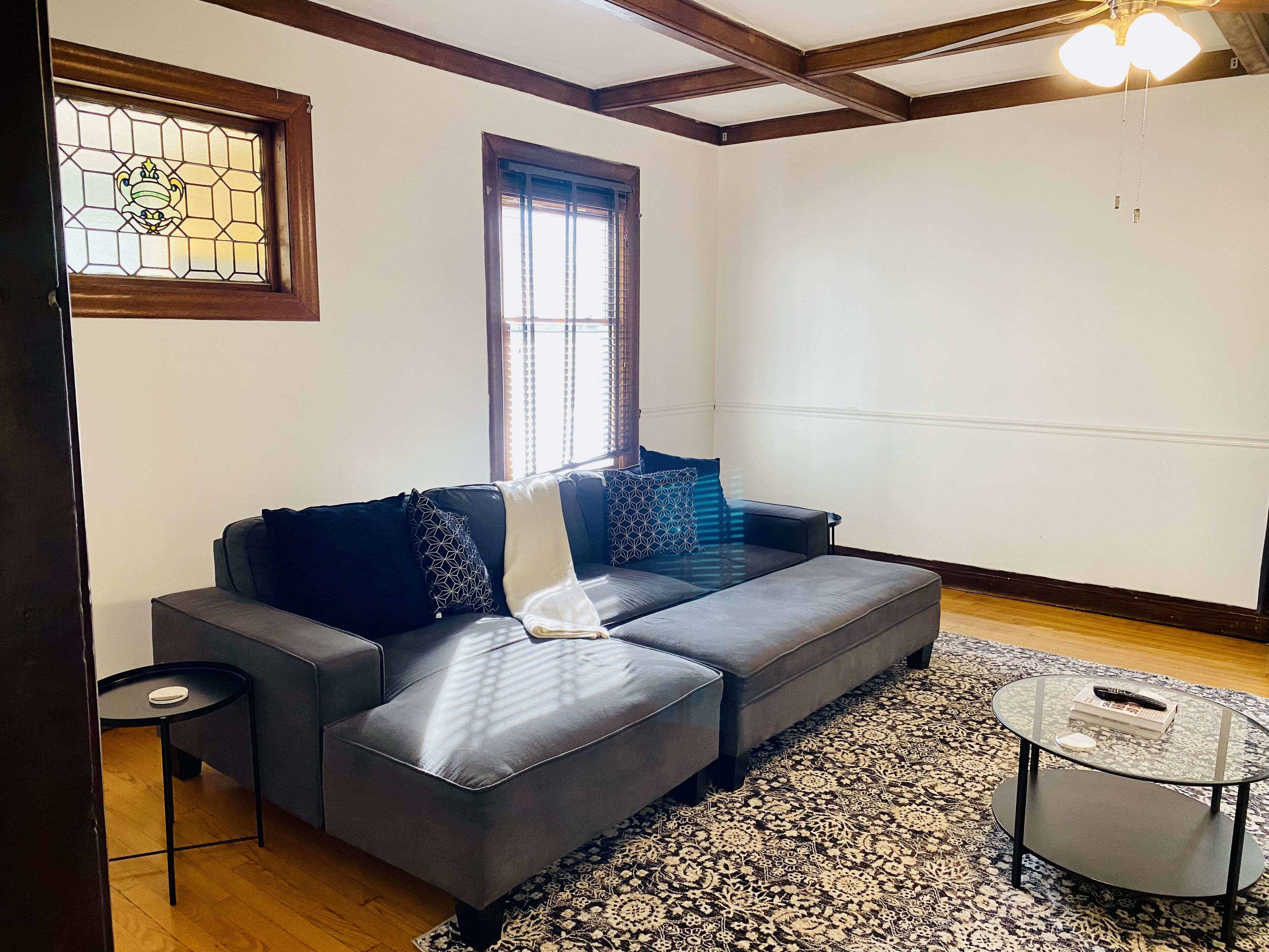 A modern living room with a grey sofa, patterned cushions, a glass coffee table, and a patterned rug.