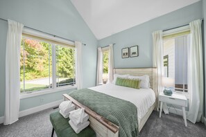 A bedroom with a large bed, a bedside table with a lamp, and a view of trees through the windows.