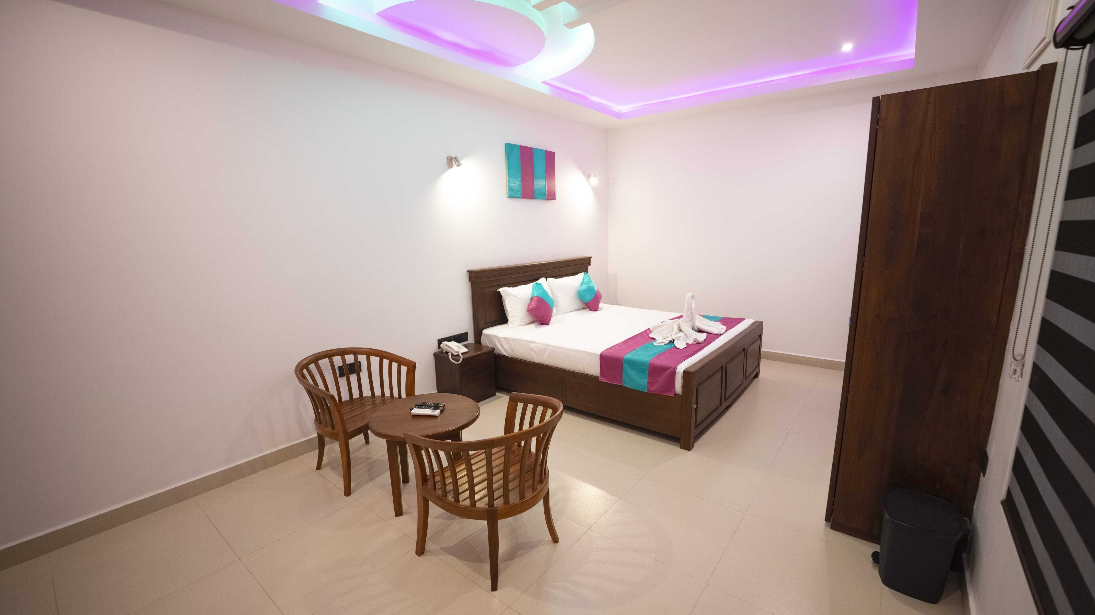 Una habitación de hotel con una cama, dos sillas de madera, una mesita pequeña, una mesita de noche con un teléfono y una obra de arte colgada en la pared.
