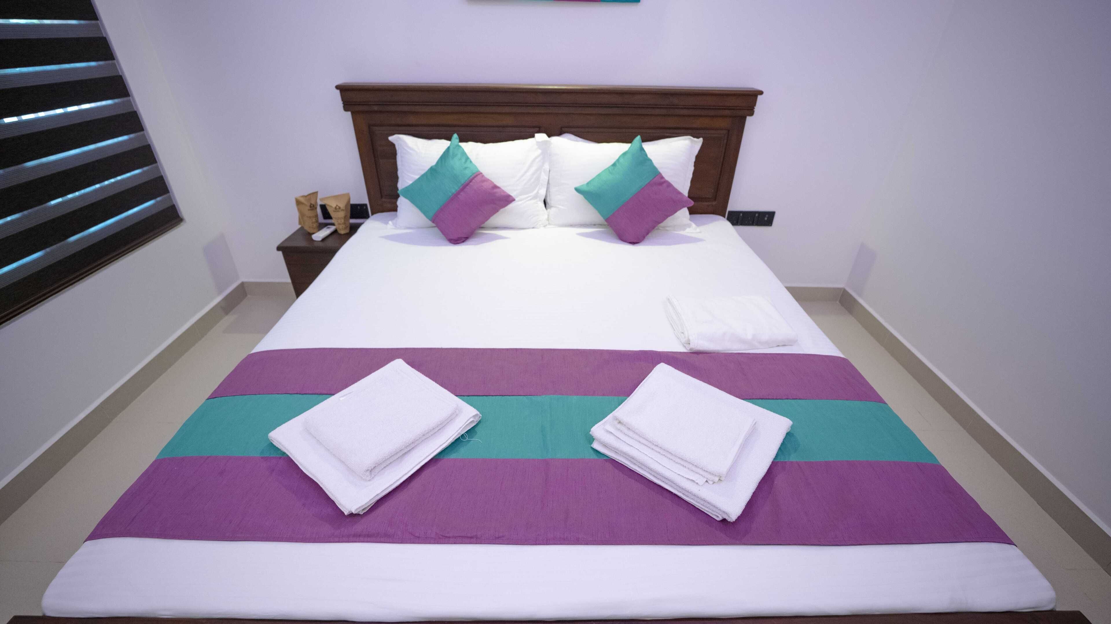 Una cama bien hecha con ropa de cama blanca y morada, dos toallas dobladas y un cabecero de madera.