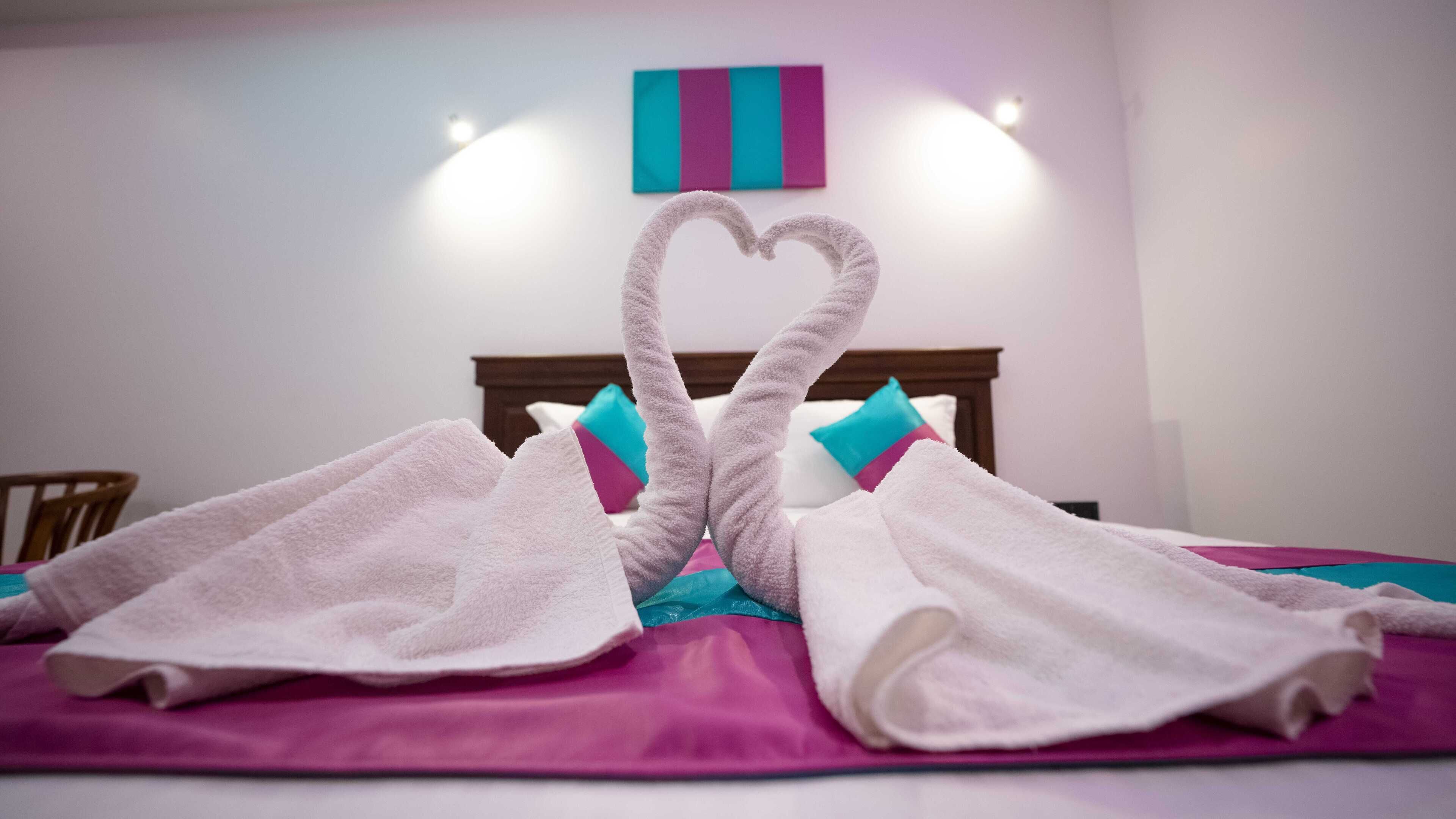 A bed with two towels arranged in a heart shape, a wooden headboard, and a colorful pillow.