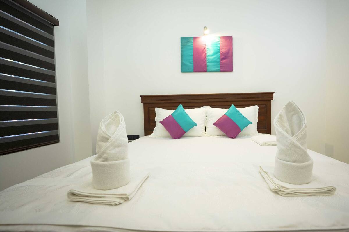 A neatly made bed with white linens, two pillows with a teal and purple design, and two folded towels on either side.