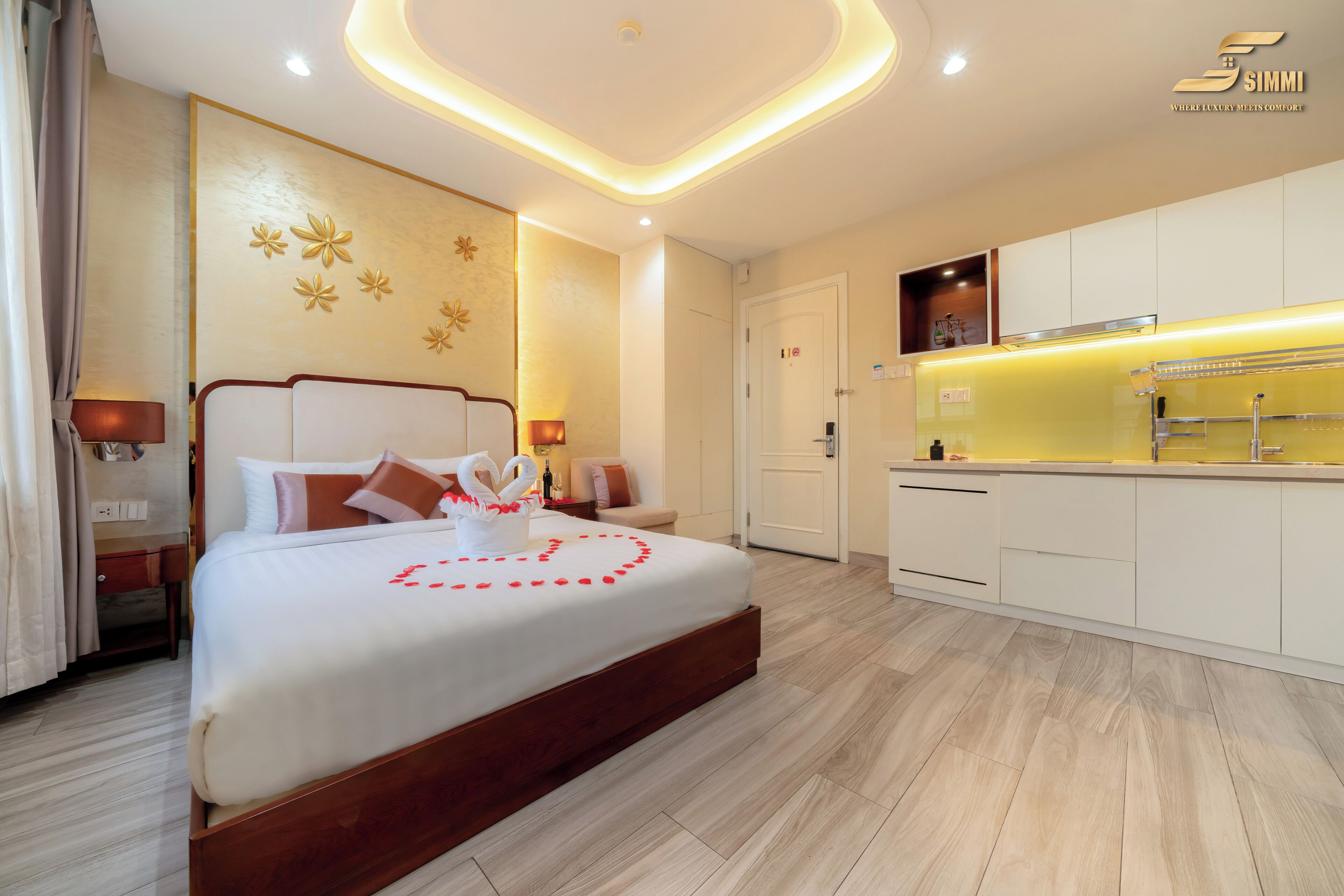 A modern hotel room with a large bed, a small desk, a vanity, and a kitchenette.