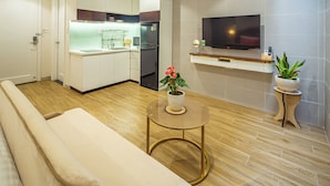 A modern living room with a sofa, a small round table with a vase, a TV on a stand, and a kitchen area with cabinets and a refrigerator.