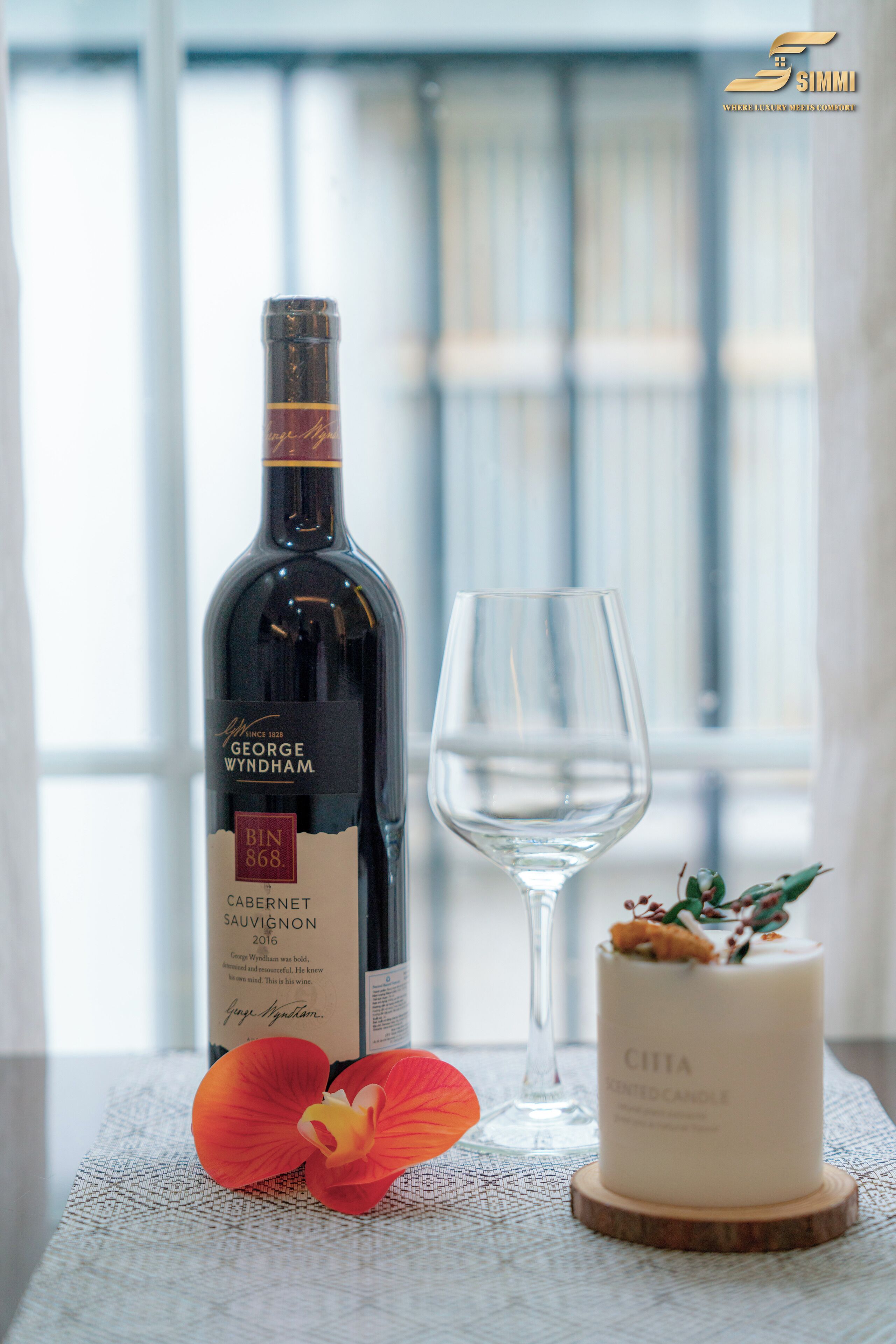 A bottle of George Wyndham Cabernet Sauvignon 2016, a glass of wine, a candle, and a red orchid on a table.