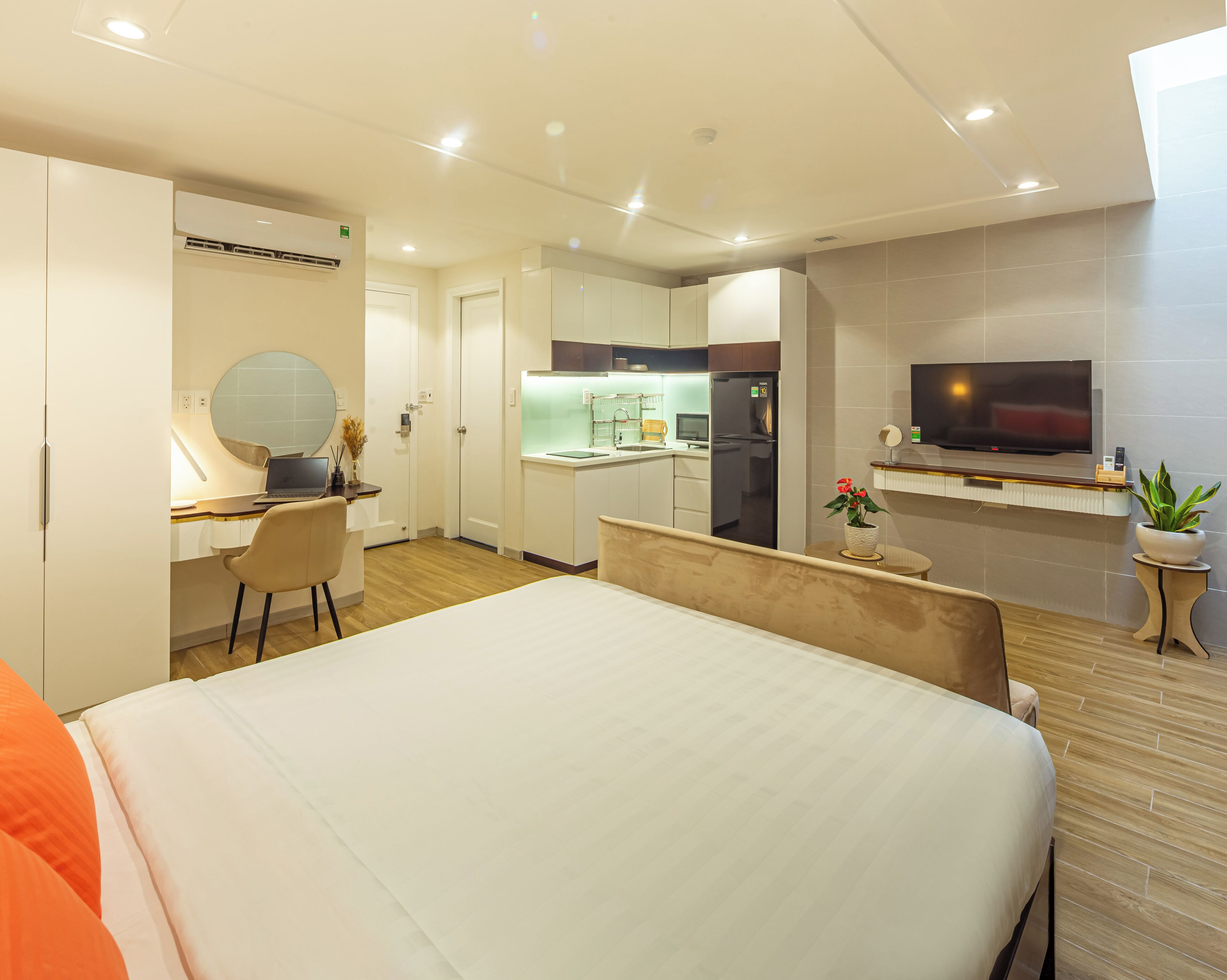 A modern hotel room with a large bed, a desk with a chair, a kitchenette, a TV, and a bathroom.