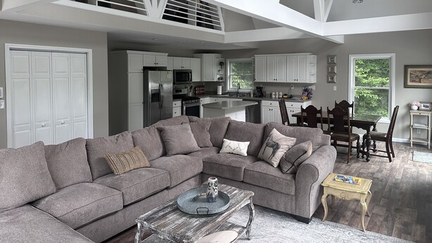 A spacious living room with a grey sectional sofa, a coffee table, and a dining area with a wooden table and chairs. A kitchen with white cabinets and stainless steel appliances is visible in the background. - Spacious home, 12 miles from Nubs Nob, Harbor Springs surrounded by forest. (Alanson)