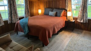 Un dormitorio con techo de paneles de madera, una cama grande con colcha roja, dos lámparas de noche y vistas a un césped verde a través de las ventanas.