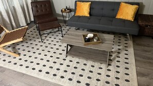Un salón con un sillón de cuero marrón, un sofá gris con almohadones amarillos, una mesa de centro de madera con adornos y una alfombra de lunares.