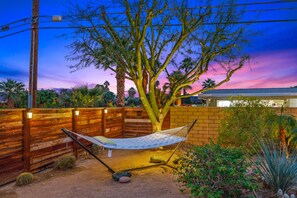 A hammock under a tree in a backyard with a wooden fence, a brick wall, and various plants. - Casita Cerritos  (Palm Springs)