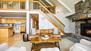 A spacious living area with a wooden staircase, a stone fireplace, and a kitchen in the background. - Luxe Mountainside Home with Panoramic Valley Views (Quechee)