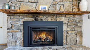 A stone fireplace with a black frame, a shelf above it, and a framed picture. - Luxe Mountainside Home with Panoramic Valley Views (Quechee)
