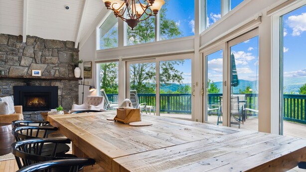 A spacious dining area with a long wooden table, stone fireplace, and large windows offering a view of the outdoors. - Luxe Mountainside Home with Panoramic Valley Views (Quechee)