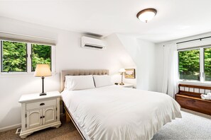 Ein Schlafzimmer mit Bett, Nachttischen, einer Lampe, einer Klimaanlage und einem Fenster mit Blick auf Grünflächen.
