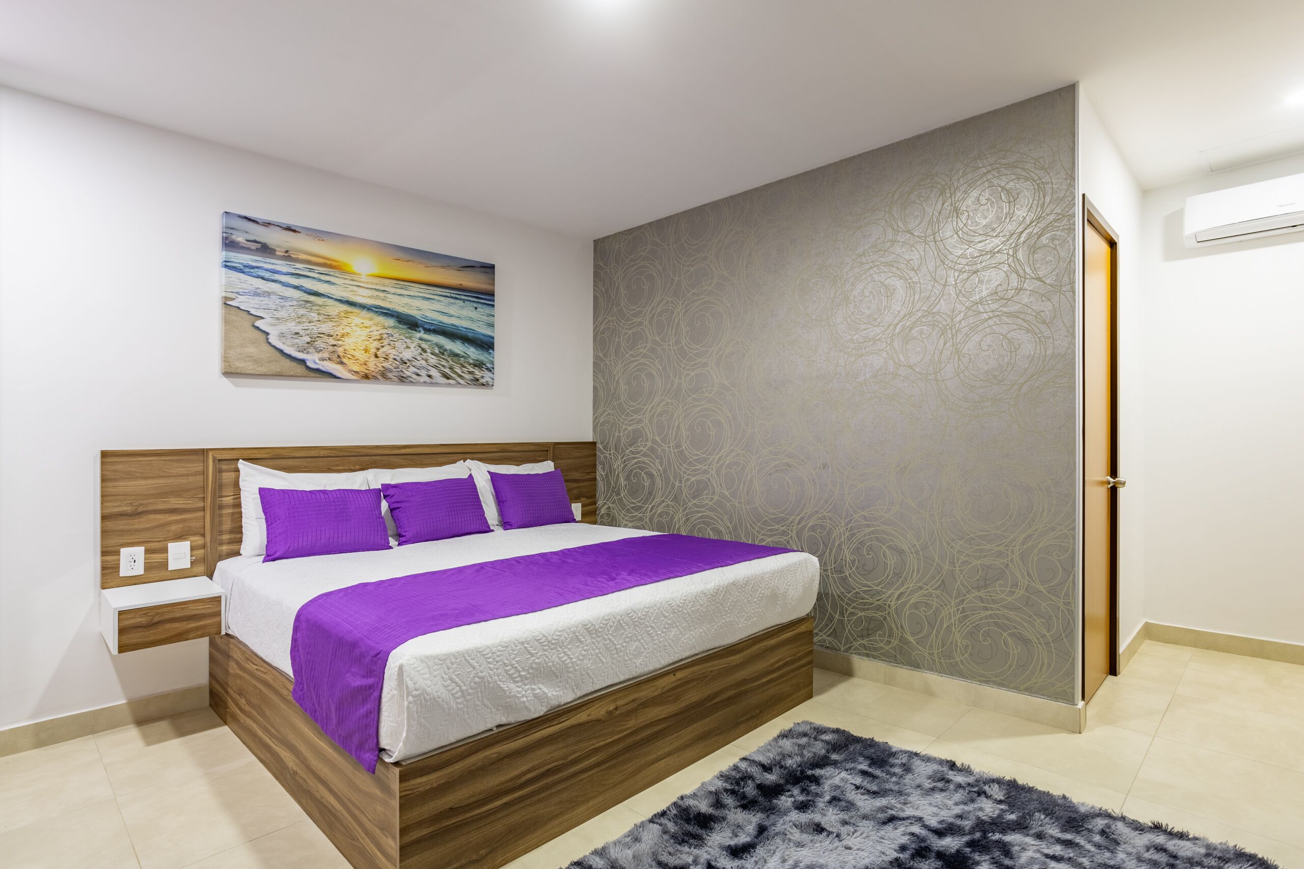Un dormitorio con cama de madera, ropa de cama blanca, un plaid púrpura, una alfombra gris, un cuadro colgado en la pared y un papel tapiz estampado.