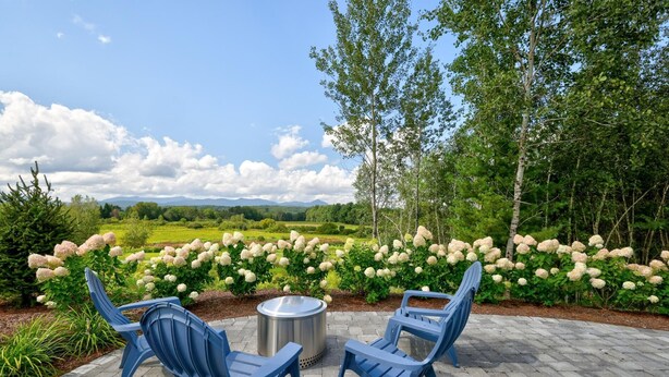 An outdoor seating area with two blue chairs and a metal table, surrounded by a garden with hydrangea flowers and trees. - Picturesque Farmhouse Retreat Minutes from Stowe (Morrisville)