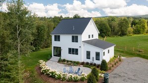 A two-story house with a dark roof, surrounded by a well-maintained lawn and a flower bed. - Picturesque Farmhouse Retreat Minutes from Stowe (Morrisville)