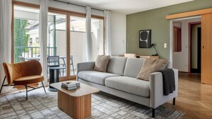 Un salon comprenant un canapé, un fauteuil et une table basse. Un balcon avec des meubles d’extérieur est visible à travers les portes vitrées.