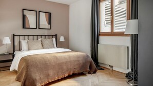 Une chambre à coucher comprenant un lit, deux tables de chevet, une fenêtre avec des rideaux, un radiateur et deux cadres photos accrochés au mur.