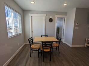 Un coin repas comprenant une table rectangulaire et quatre chaises, une fenêtre avec des stores, une porte et une horloge fixée au mur.