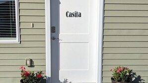 A white door with the name "Casita" on it, flanked by two potted plants with red flowers, and a doormat with a floral design.