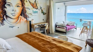 Una habitación con cama, escritorio, balcón con vista al mar y un mural en la pared.