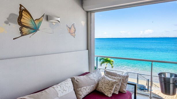 Una habitación con un amplio ventanal que da a la playa, un sofá con cojines estampados y cuadros de mariposas en la pared.